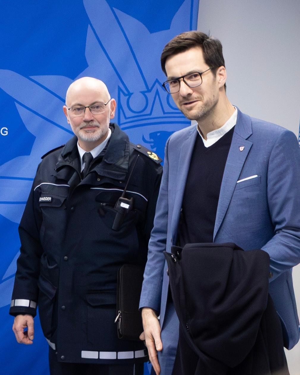 Dieser Alternativtext wurde KI-generiert: Zwei Personen in formeller Kleidung stehen vor einem blauen Hintergrund mit einem weißen Emblem. Eine Person trägt eine Uniform, die andere einen Anzug und eine Jacke.