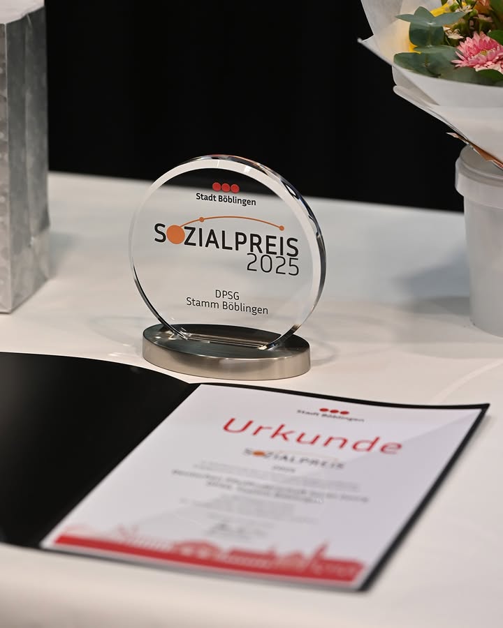 Dieser Alternativtext wurde KI-generiert: Eine Glasauszeichnung und eine Urkunde liegen nebeneinander auf einer weißen Oberfläche. Der Preis trägt die Aufschrift "Sozialpreis 2025" und weitere Details.