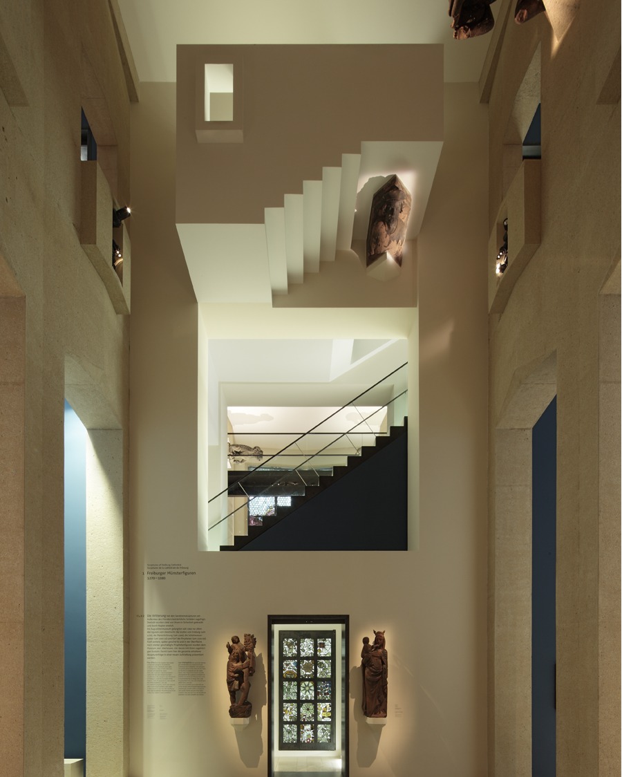 Dieser Alternativtext wurde KI-generiert: Ein Innenraum mit einer Treppe, die durch mehrere Stockwerke führt. An den Wänden befinden sich Kunstwerke und Informationstafeln.