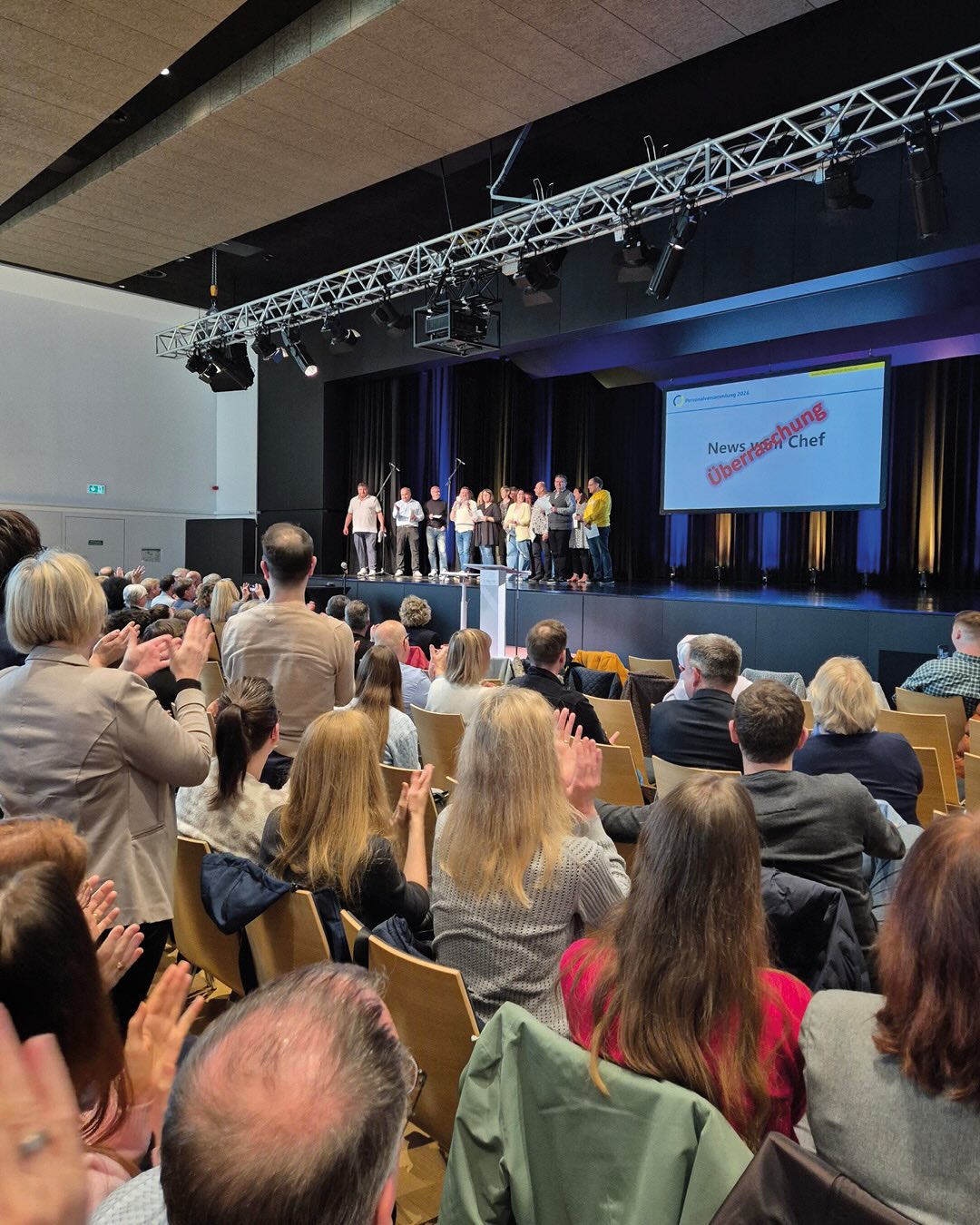 Dieser Alternativtext wurde KI-generiert: Eine Gruppe von Personen steht auf einer Bühne vor Publikum. Im Hintergrund ist ein Raum mit Stühlen und einem Banner zu sehen.