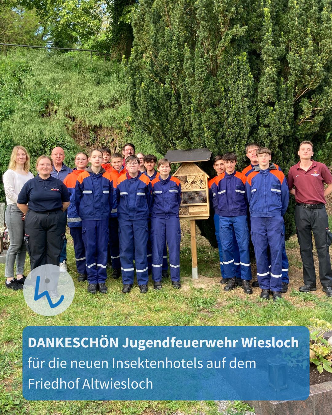 Gruppe von Jugendlichen und Erwachsenen in blau-orangen Arbeitsanzügen steht vor einem Insektenhotel im Freien; Text am Bildrand dankt der Jugendfeuerwehr Wiesloch für die neuen Insektenhotels auf dem Friedhof Altwiesloch. (KI-Alternativtext)
