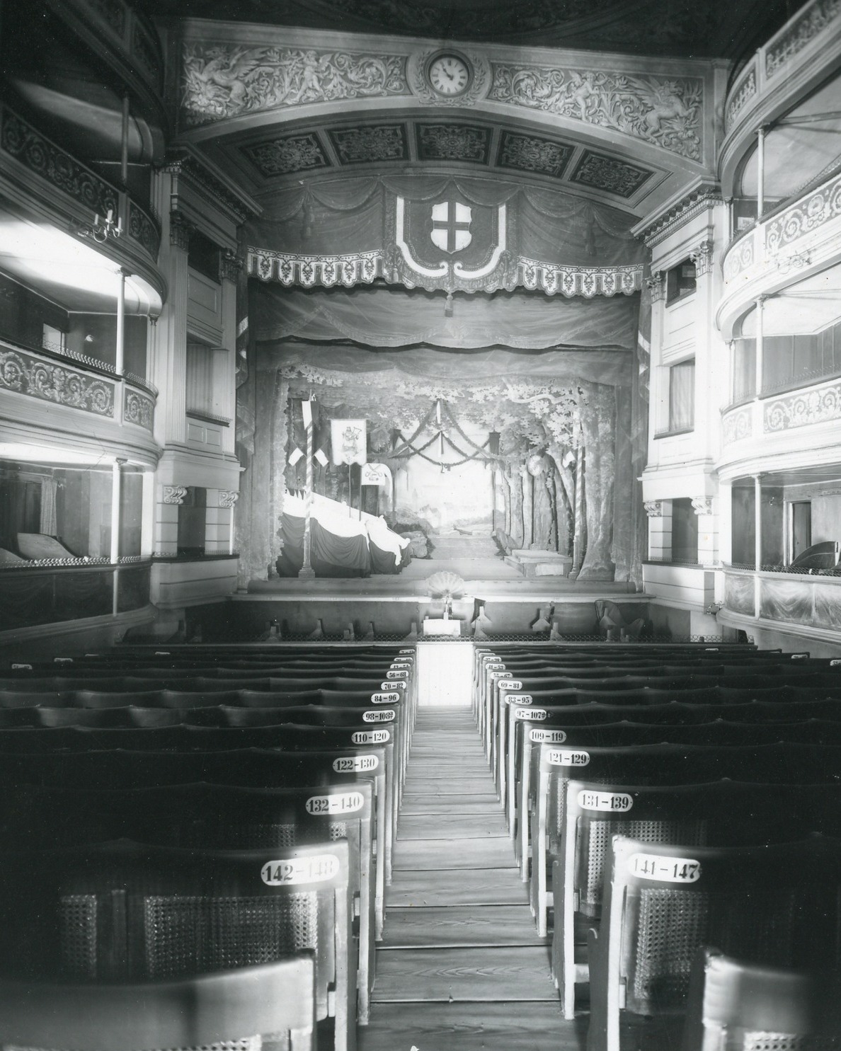 Dieser Alternativtext wurde KI-generiert: Schwarz-Weiß-Fotografie eines Innenraums mit Reihen von Sitzplätzen, die auf eine Bühne zulaufen. Ein dekorativer Bogen und ein Uhrenmotiv sind ebenfalls zu sehen.