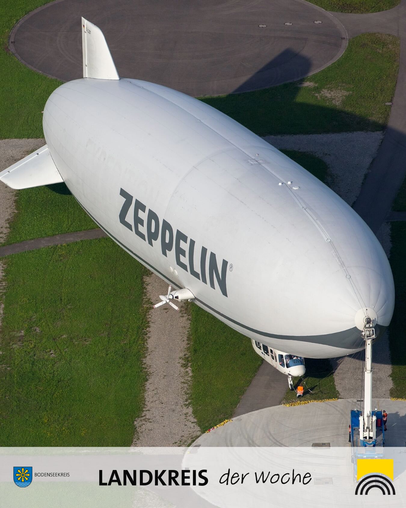 Ein weißes Zeppelin-Luftschiff mit der Aufschrift „ZEPPPELIN“ ist am Boden vertäut, umgeben von Grünflächen und einem Betonplatz. Unten im Bild steht „LANDKREIS der Woche“ neben dem Logo des Bodenseekreises. (KI-Alternativtext)