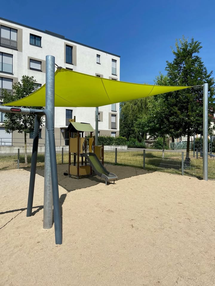 Ein Spielplatz mit Sandboden, der von einem großen, gelben Sonnensegel beschattet wird. Unter dem Segel befindet sich eine kleine Holzspielstruktur mit einer Rutsche. Im Hintergrund steht ein weißes Mehrfamilienhaus neben einigen Bäumen. (KI-Alternativtext)