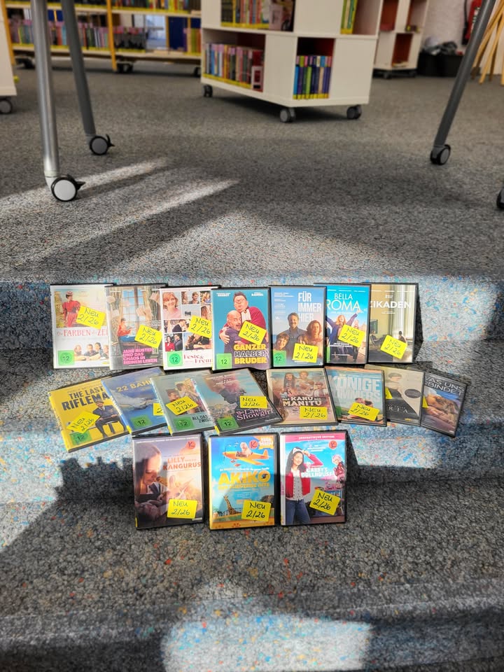 Dieser Alternativtext wurde KI-generiert: Eine Auswahl an Medien liegt auf einem blauen Teppichboden vor einem Bücherregal. Die Medien scheinen Zeitschriften und DVDs zu sein, einige mit gelben Aufklebern versehen.