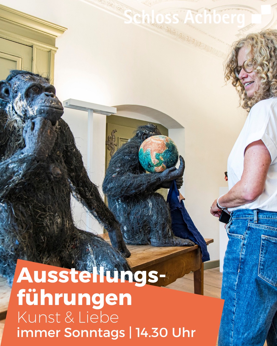 Ein helles Innenraumbild zeigt zwei skulpturale affenaehnliche Figuren, dazwischen steht eine Person, und einen orangefarbenen Block mit Ausstellungsdetails.
