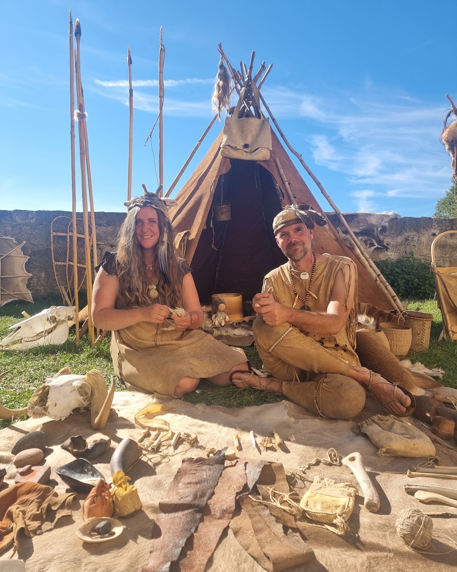 Dieser Alternativtext wurde KI-generiert: Ein Zelt und verschiedene Objekte stehen vor einer Steinmauer. Zwei Personen sitzen davor und tragen historische Kleidung.