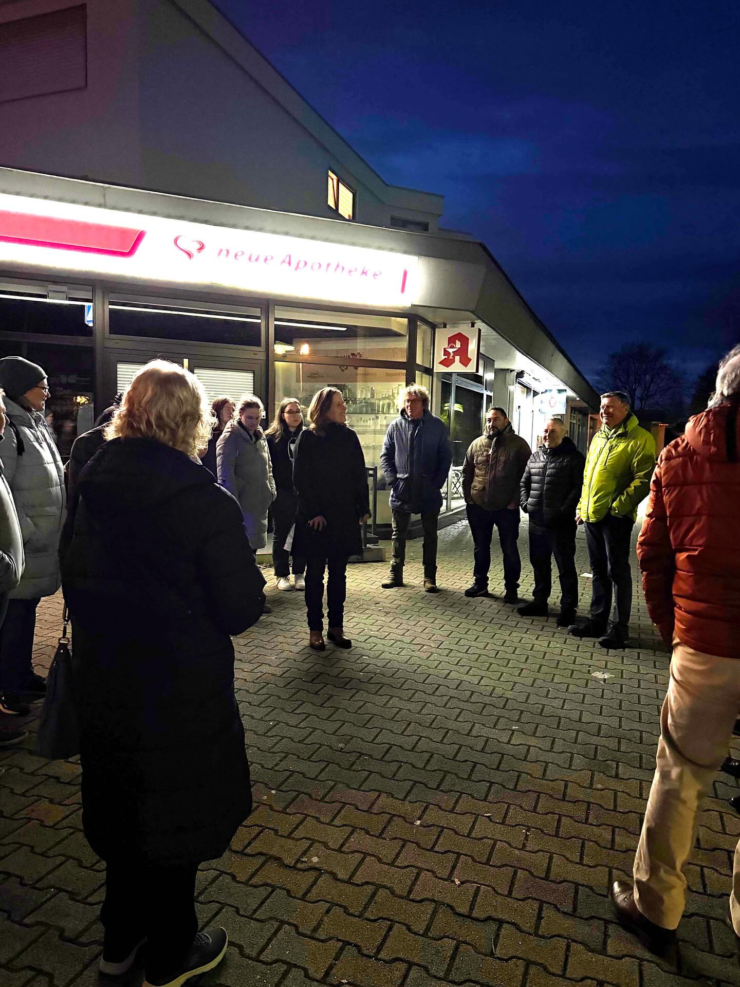 Dieser Alternativtext wurde KI-generiert: Eine Gruppe von Menschen steht vor dem Eingang eines Gebäudes mit einem roten Schild. Das Foto ist bei Nacht aufgenommen und zeigt eine Szene im Freien.