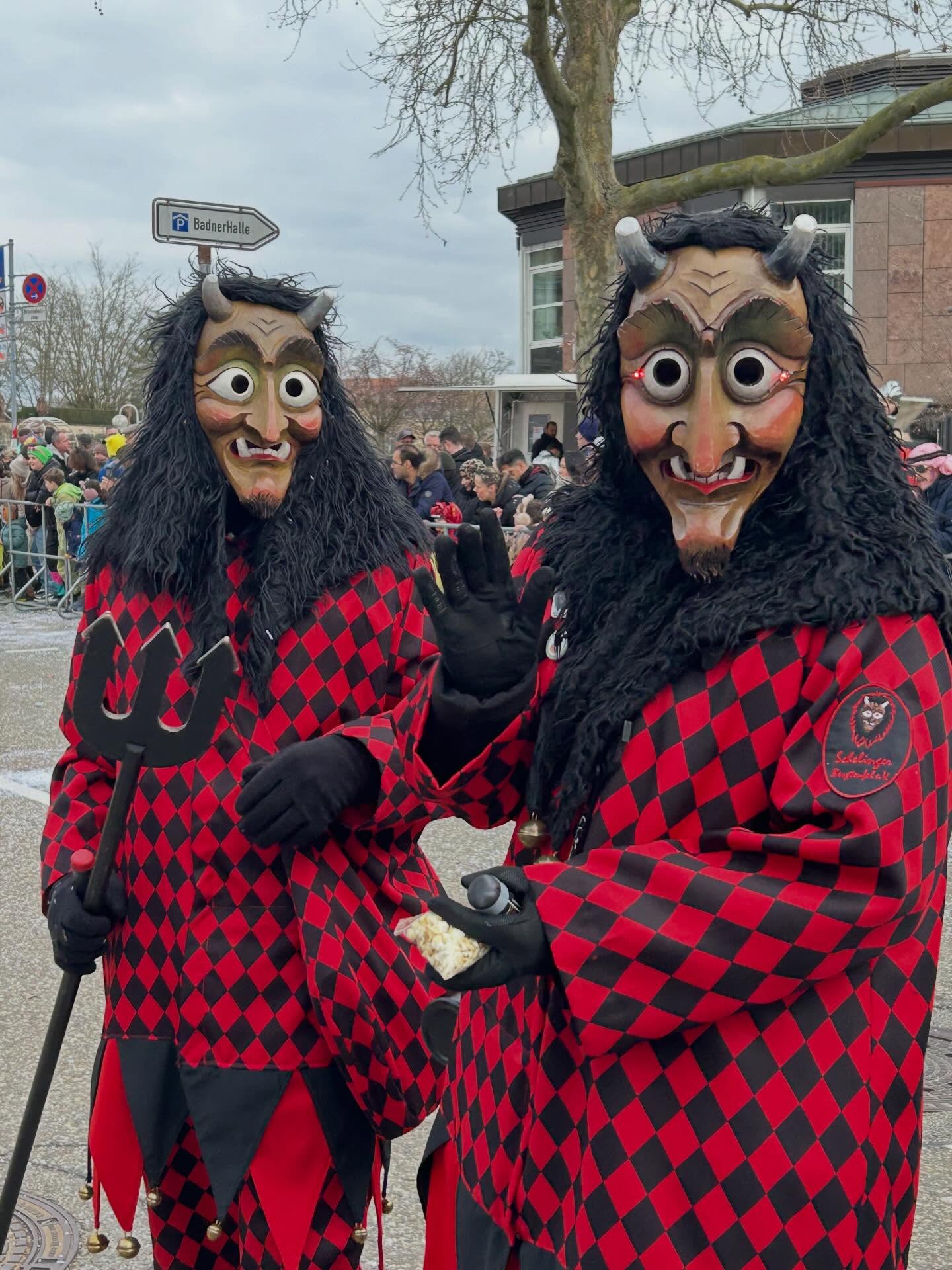 Dieser Alternativtext wurde KI-generiert: Zwei Figuren mit auffälligen, roten Kostümen und markanten Masken stehen im Freien. Im Hintergrund sind Menschen und Gebäude zu sehen.