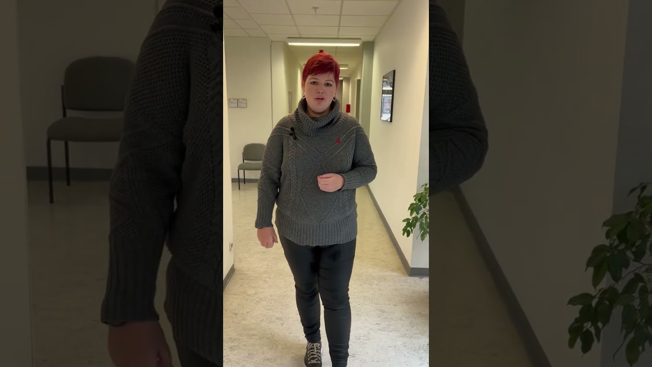 Dieser Alternativtext wurde KI-generiert: Eine Person steht in einem Flur, der mit mehreren Türen versehen ist. Im Hintergrund sind Stühle und Pflanzen erkennbar.