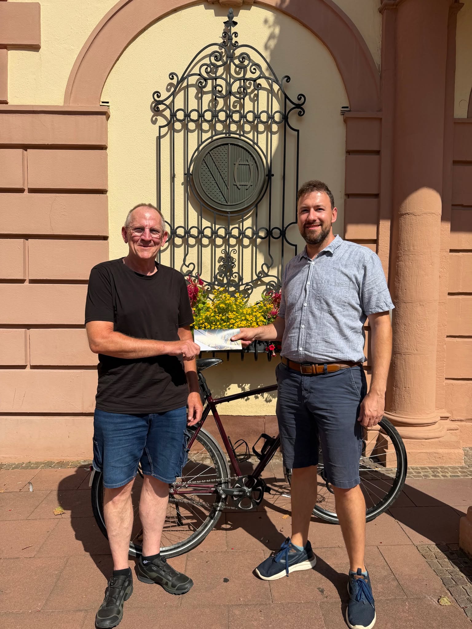 Zwei Personen, eine in einem schwarzen T-Shirt und die andere in einem hellblauen Hemd, stehen vor einem Gebäude mit einem großen Gitterfenster und halten gemeinsam ein Blatt Papier. Zwischen ihnen steht ein Fahrrad. (KI-Alternativtext)