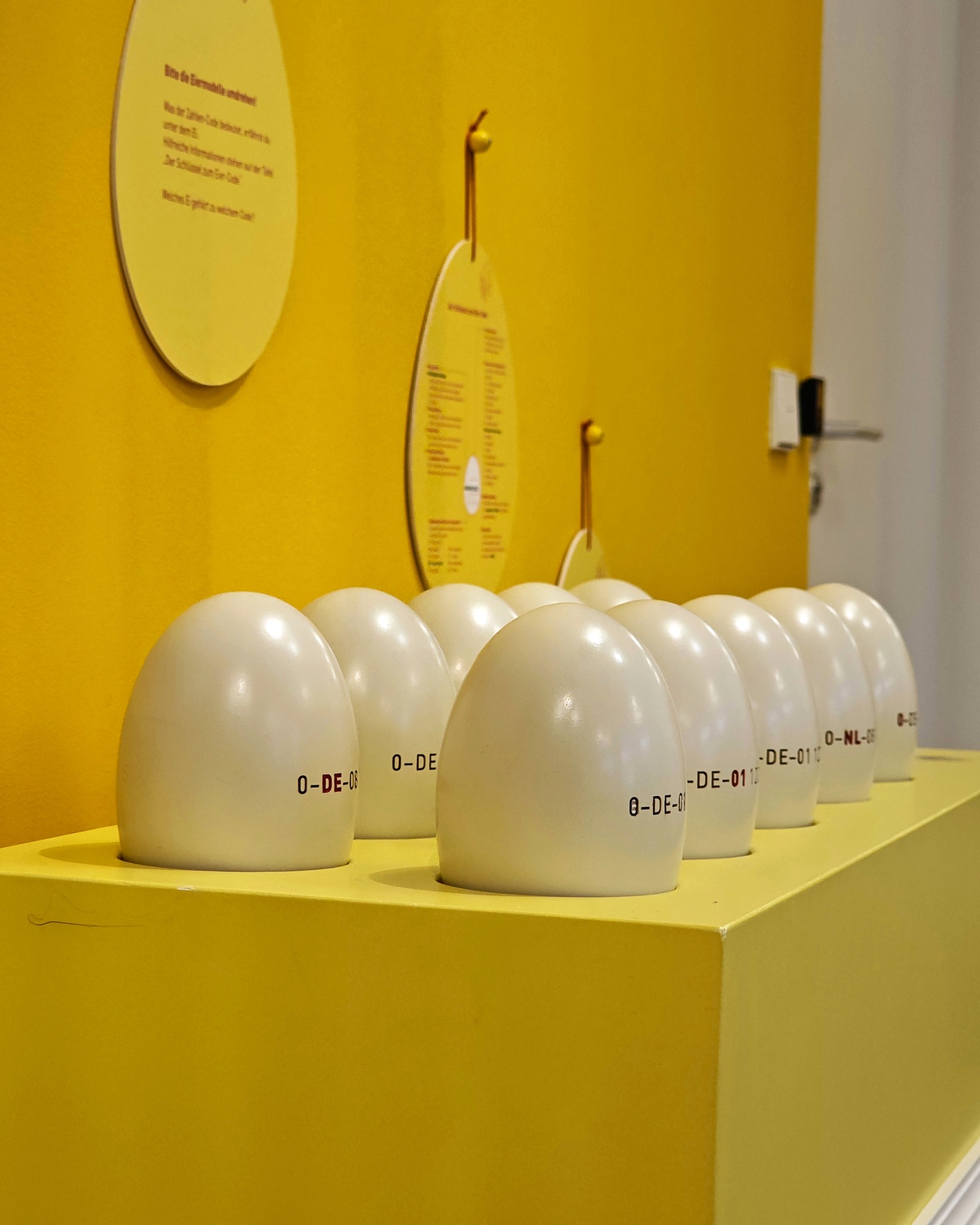 Dieser Alternativtext wurde KI-generiert: Eine Anordnung von weißen, ovalen Objekten steht vor einer gelben Wand. Die Objekte haben Beschriftungen und sind in einer Reihe auf einem gelben Podest platziert.