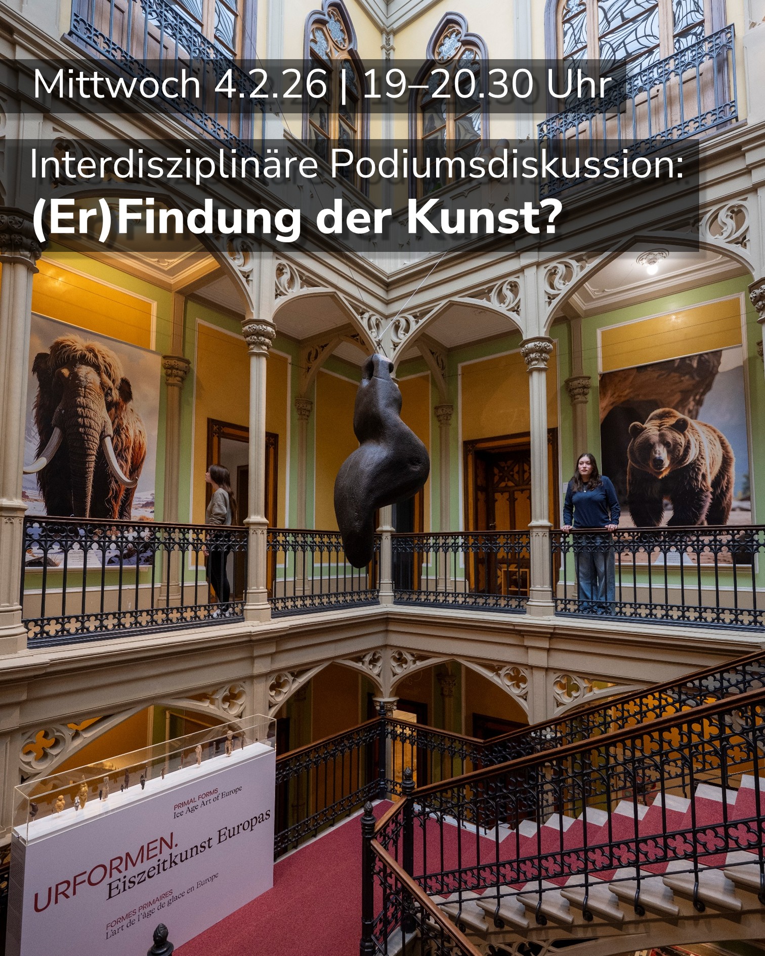 Dieser Alternativtext wurde KI-generiert: Ein Innenraum mit einer Wendeltreppe und Säulen. Im Hintergrund sind Kunstwerke und ein Stand mit Informationen zu sehen.