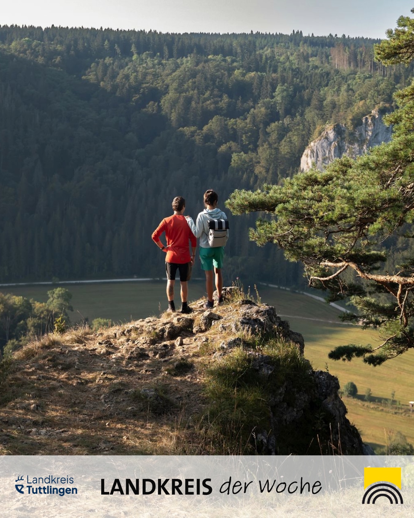 Dieser Alternativtext wurde KI-generiert: Zwei Personen stehen mit dem Rücken zum Betrachter auf einer Anhöhe und blicken auf eine bewaldete Landschaft. Das Bild ist farbig und zeigt eine Aussicht in der Natur.