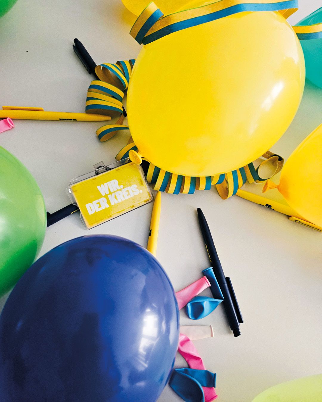 Dieser Alternativtext wurde KI-generiert: Mehrere Luftballons in verschiedenen Farben liegen neben einer Girlande und Stiften. Das Bild ist farbig und zeigt eine festliche Dekoration.