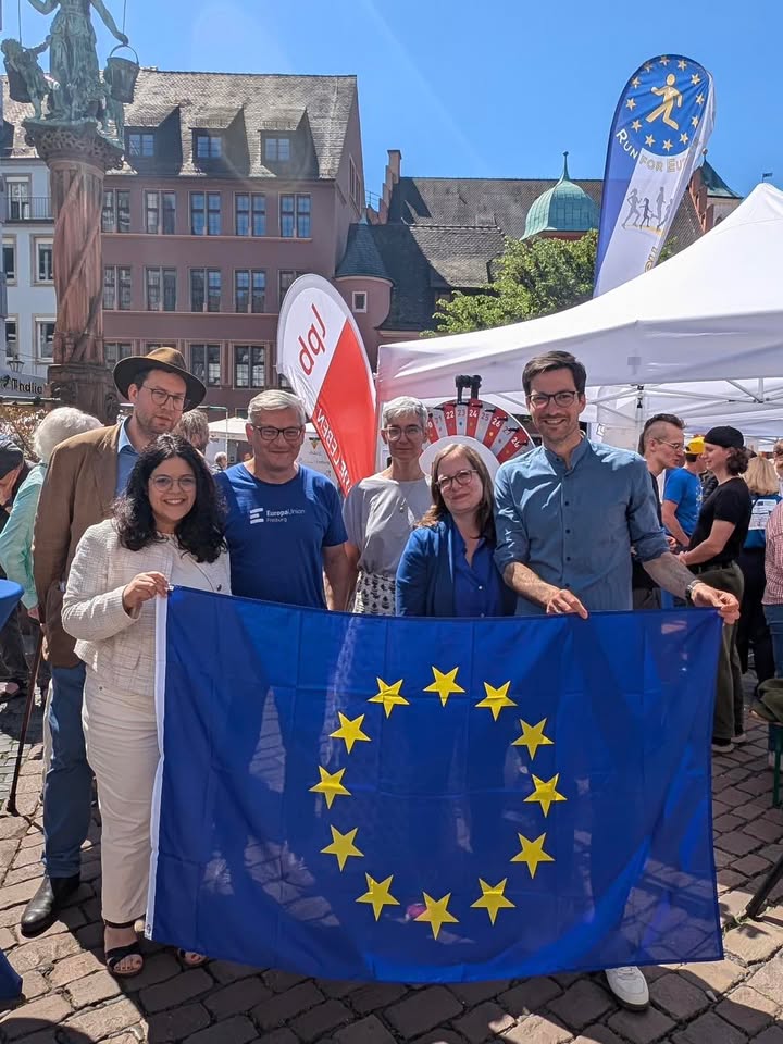Dieser Alternativtext wurde KI-generiert: Eine Gruppe von Menschen steht im Freien vor Gebäuden mit einer Flagge mit Sternen und einem Banner. Im Hintergrund ist eine blaue und gelbe Flagge zu sehen.