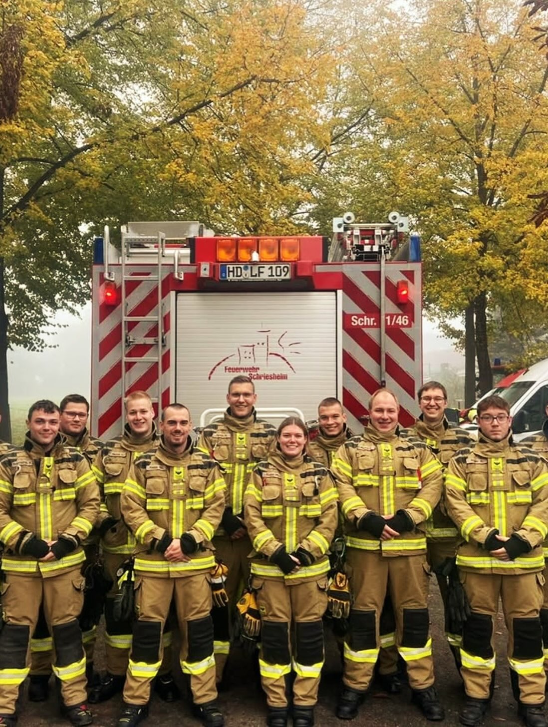 Dieser Alternativtext wurde KI-generiert: Eine Gruppe von Menschen in Uniformen steht vor einem roten Feuerwehrauto. Im Hintergrund sind Bäume mit herbstlichen Blättern zu sehen.