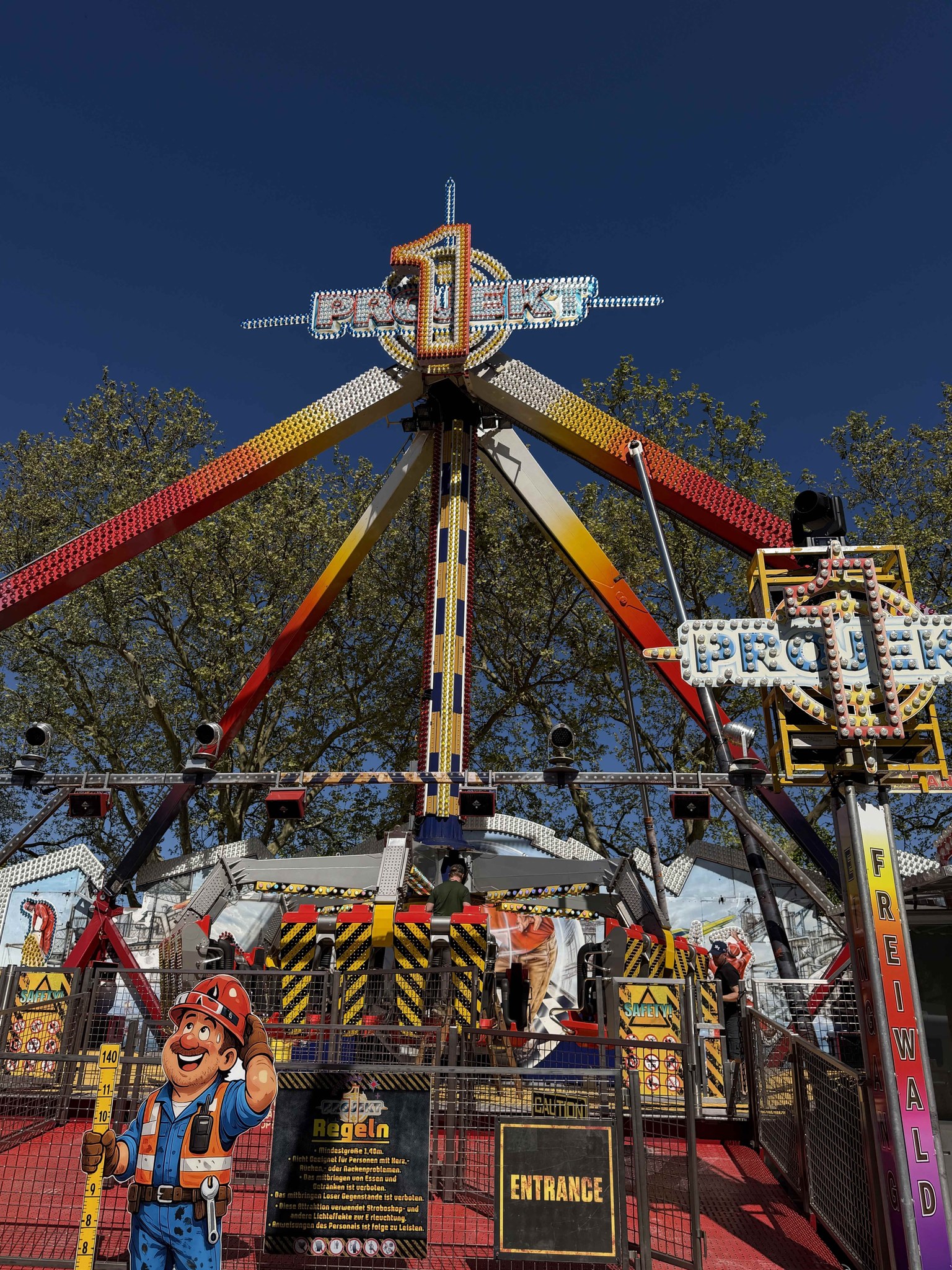 Dieser Alternativtext wurde KI-generiert: Ein buntes Fahrgeschäft auf einem Jahrmarkt, umgeben von Bäumen unter einem blauen Himmel. Das Bild ist farbig und zeigt verschiedene Details der Attraktion und der Umgebung.
