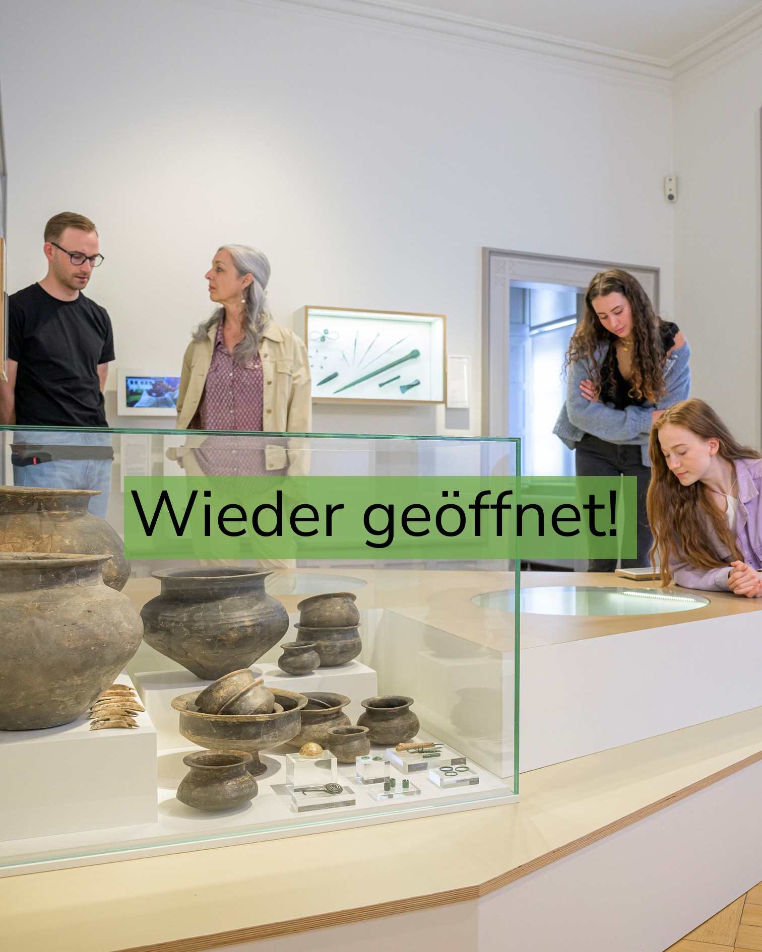 Vier Personen betrachten in einem Museum Ausstellungsstücke in einer Glasvitrine mit antiken Keramikgefäßen und kleinen Artefakten. Ein grüner Textbalken überlagert das Bild mit der Aufschrift "Wieder geöffnet!". (KI-Alternativtext)