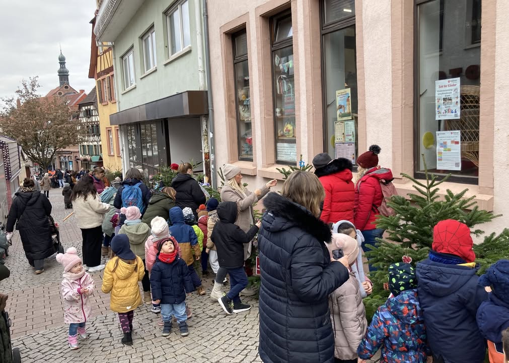Dieser Alternativtext wurde KI-generiert: Eine Gruppe von Menschen steht vor einem Geschäft auf einem Pflasterweg. Im Schaufenster sind verschiedene Gegenstände ausgestellt und ein kleiner Weihnachtsbaum steht davor.