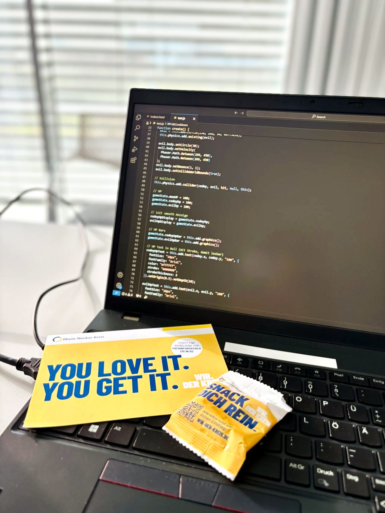 Dieser Alternativtext wurde KI-generiert: Ein geöffneter Laptop zeigt eine Code-Oberfläche. Daneben liegen verschiedene Snacks und eine Karte auf einer Tastatur.