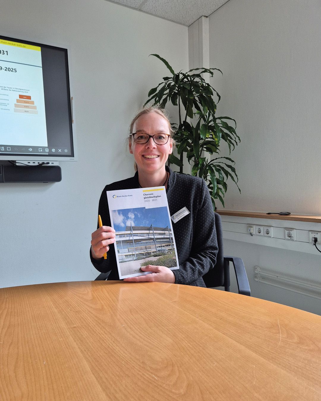 Eine Person mit Brille sitzt an einem Holztisch, hält ein Dokument mit dem Titel „Chancengleichheitsplan 2023–2027“ und einen gelben Stift. Im Hintergrund ist ein Bildschirm mit einer Präsentation und eine Pflanze zu sehen. (KI-Alternativtext)
