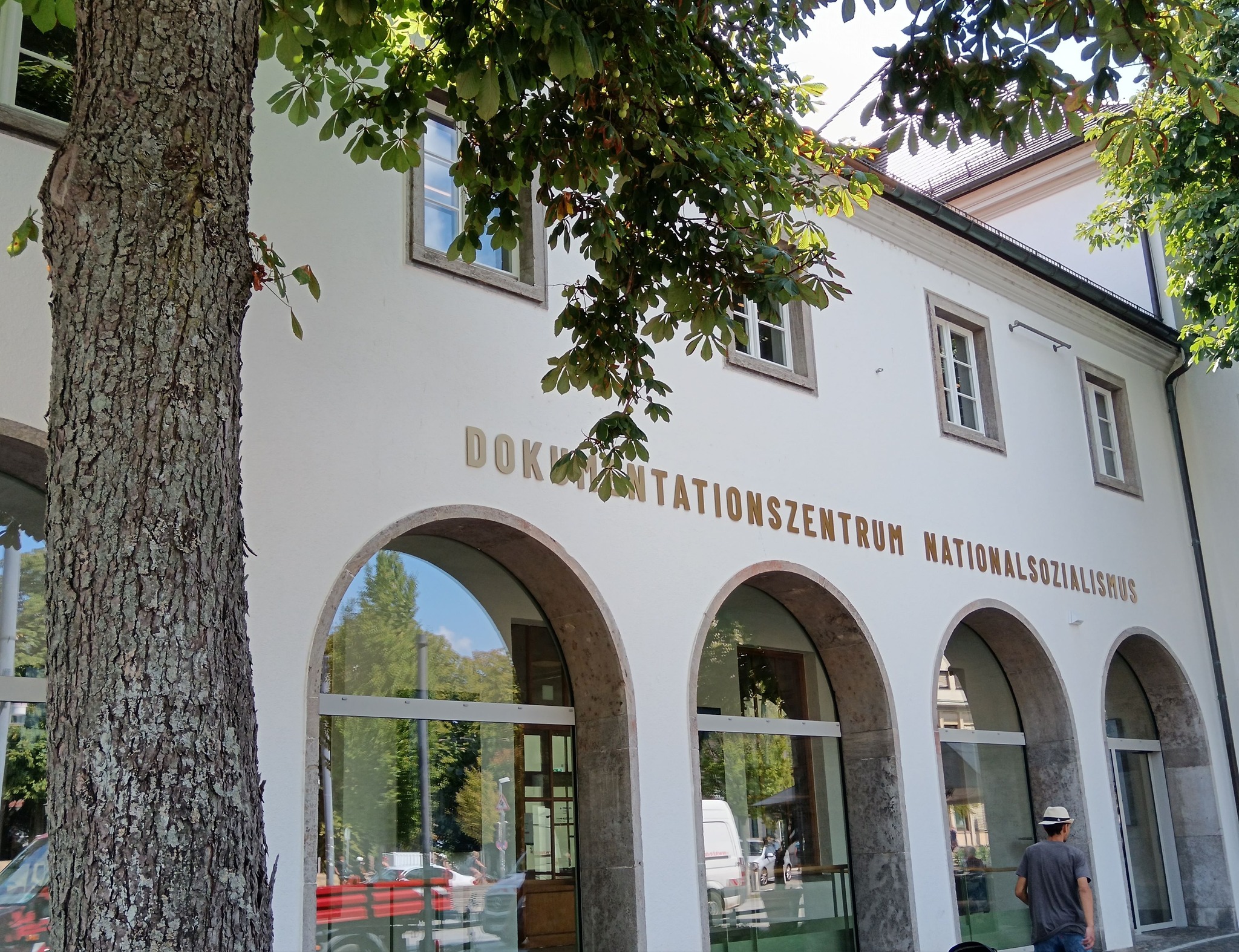 Eine weiße Gebäudefassade mit großen Bogenfenstern und der Aufschrift "DOKUMENTATIONSZENTRUM NATIONALSOZIALISMUS" in goldenen Buchstaben. Im Vordergrund links steht ein dicker Baumstamm, dessen Äste und Blätter ins Bild ragen. Rechts unten geht eine Person vorbei. (KI-Alternativtext)