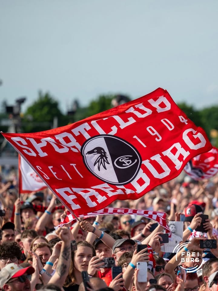 Eine Menschenmenge hält eine große rote Fahne mit dem Schriftzug „Sportclub Freiburg seit 1904“ und dem Vereinswappen hoch. Viele Personen filmen oder fotografieren mit Smartphones. (KI-Alternativtext)