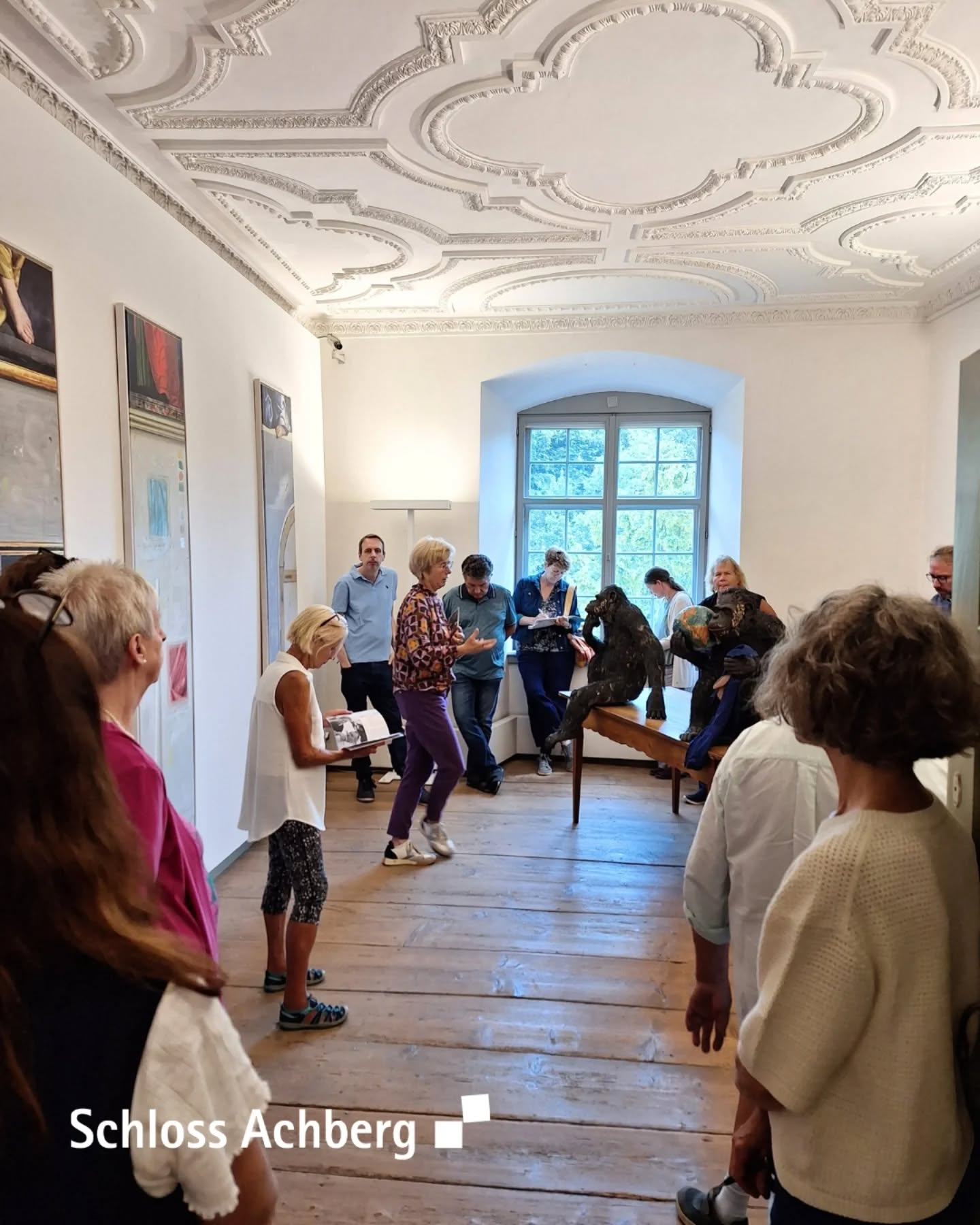 Innenansicht eines Raumes mit einer Gruppe von Menschen. An den Wänden hängen Bilder und die Decke ist mit Stuck verziert.