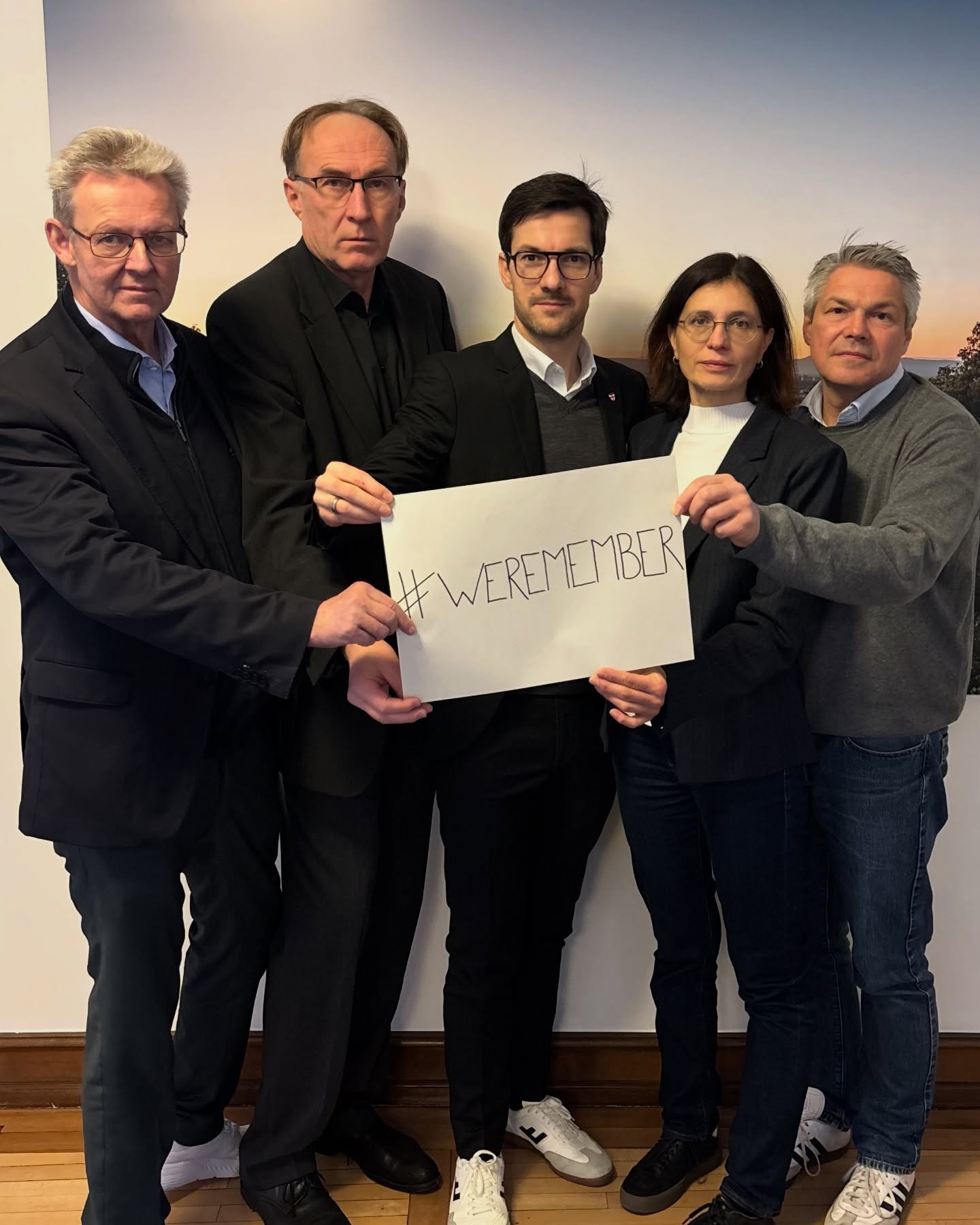 Dieser Alternativtext wurde KI-generiert: Eine Gruppe von Personen steht nebeneinander und hält ein Schild mit dem Hashtag #WEREMEMBER. Das Bild ist farbig und zeigt die Menschen vor einer hellen Wand.