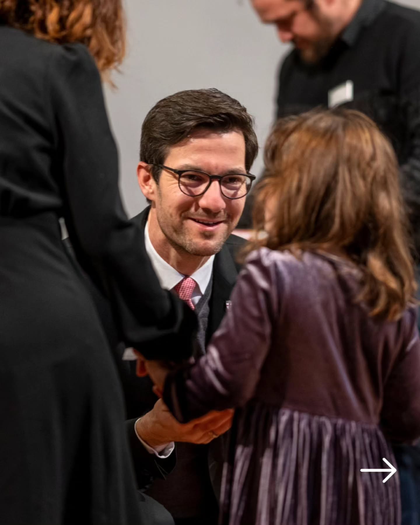 Dieser Alternativtext wurde KI-generiert: Eine Person mit Brille lächelt, während sie die Hand einer anderen Person schüttelt. Menschen stehen im Hintergrund.