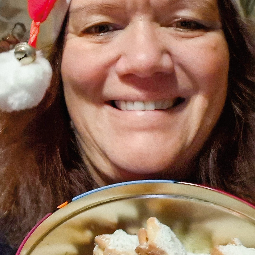 Dieser Alternativtext wurde KI-generiert: Nahaufnahme einer lächelnden Person mit roter Kopfbedeckung. Im unteren Bildrand ist ein Teil eines Tellers mit Gebäck zu sehen.