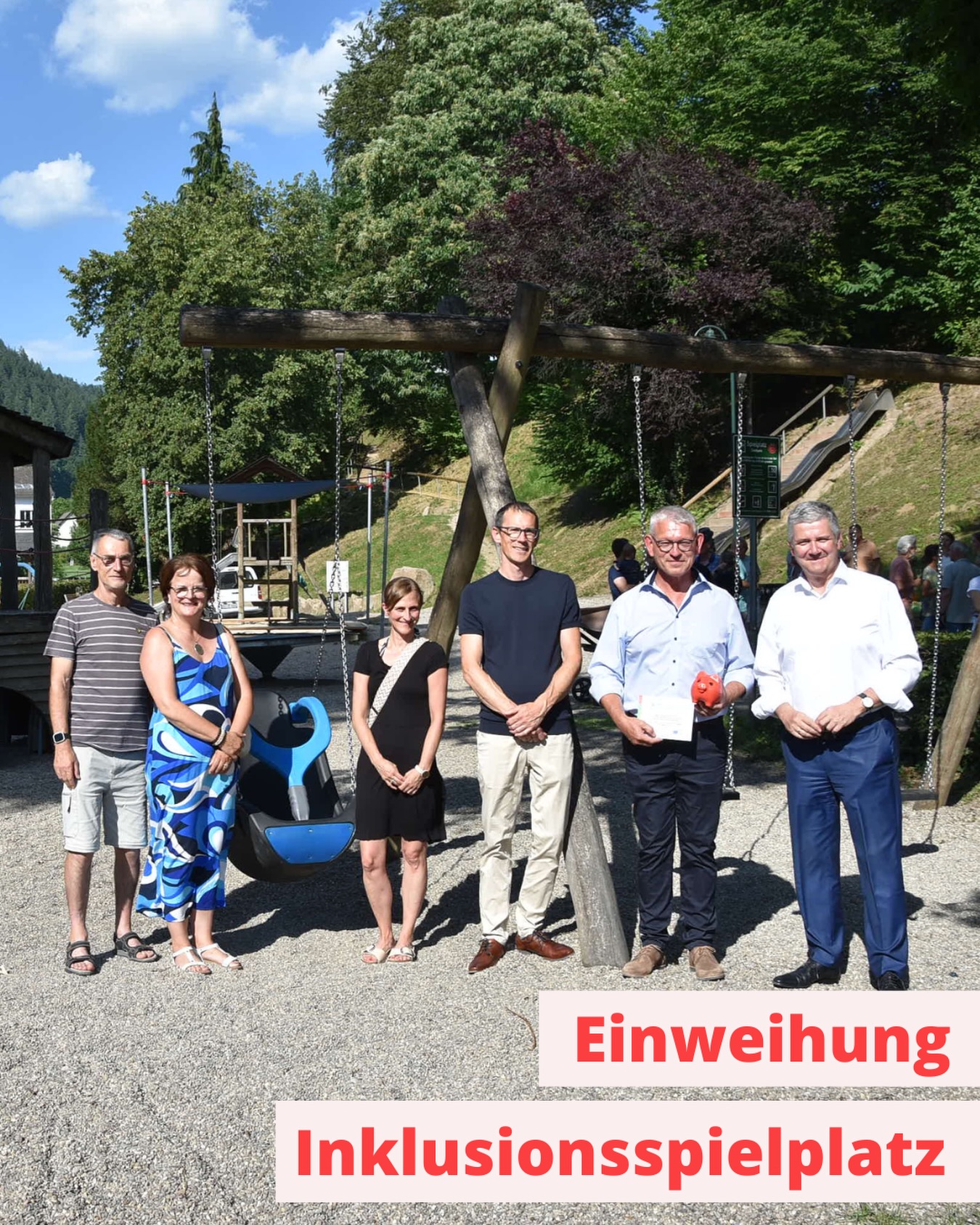 Sechs Personen stehen im Freien vor einem hölzernen Schaukelgestell auf einem Spielplatz. Eine Person hält ein Dokument und ein rotes Objekt in den Händen. Im Vordergrund steht der rote Text „Einweihung Inklusionsspielplatz“ auf einem hellen Hintergrund. (KI-Alternativtext)