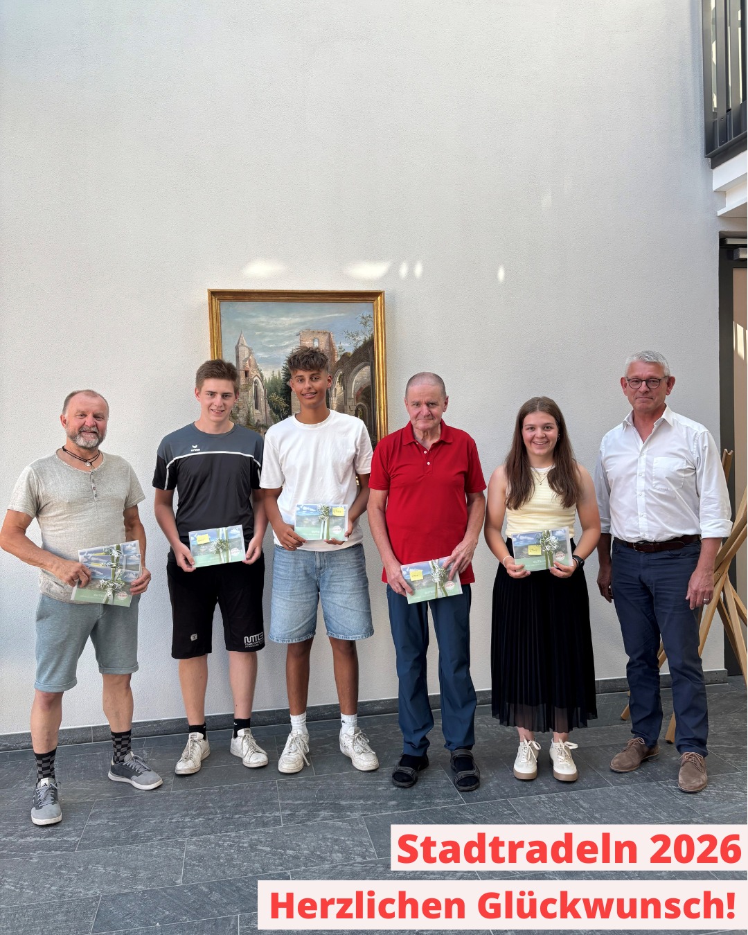 Sechs Personen stehen nebeneinander vor einer weißen Wand mit einem gerahmten Gemälde und halten jeweils ein Zertifikat in den Händen. Im unteren Bildbereich ist der rote Text „Stadtradeln 2026 Herzlichen Glückwunsch!“ eingeblendet. (KI-Alternativtext)