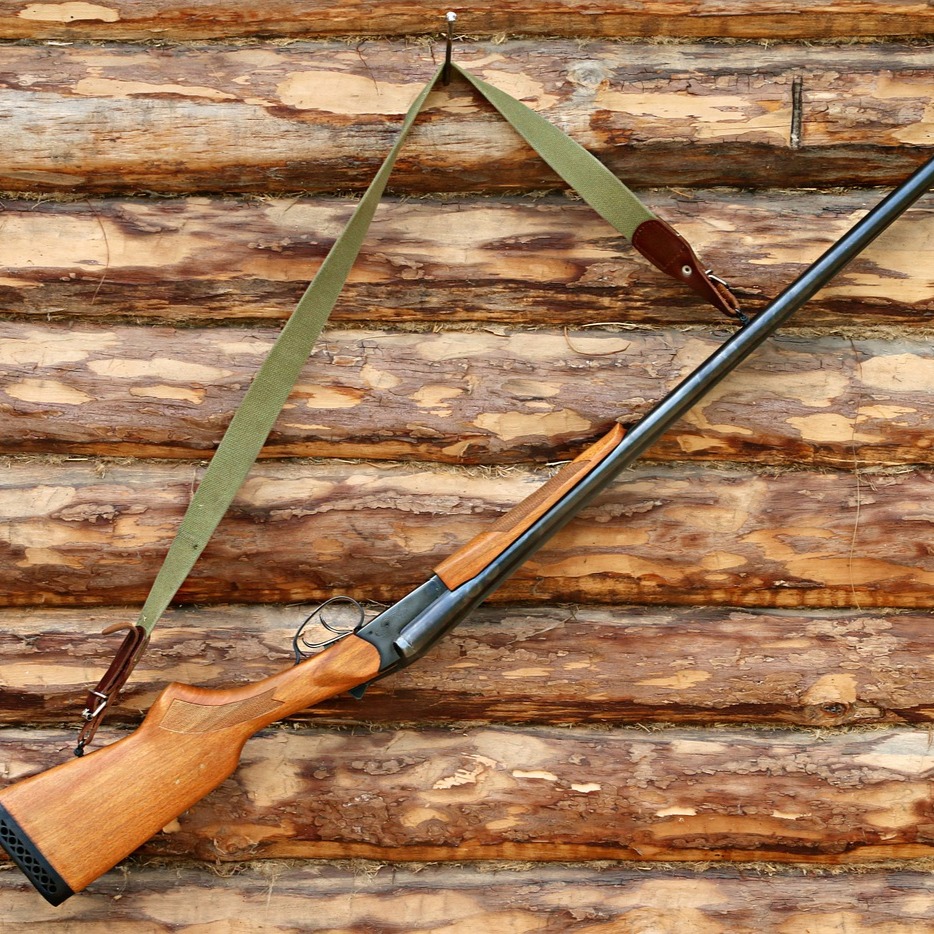 Dieser Alternativtext wurde KI-generiert: Ein Gewehr mit Tragegurt lehnt an einer Holzwand. Das Bild ist in warmen Brauntönen gehalten.