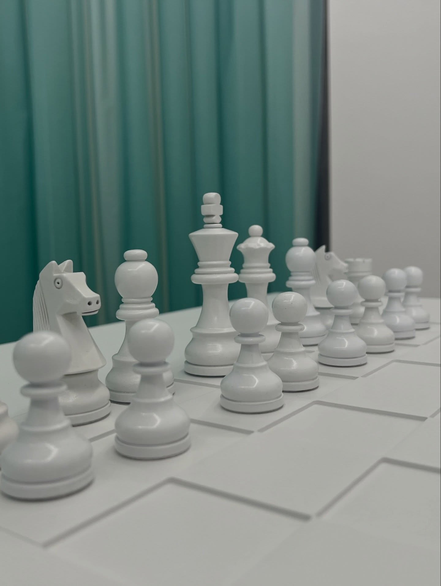 Dieser Alternativtext wurde KI-generiert: Eine Anordnung weißer Schachfiguren steht auf einem Schachbrett vor einem türkisfarbenen Hintergrund mit Vorhängen. Das Bild ist farbig.