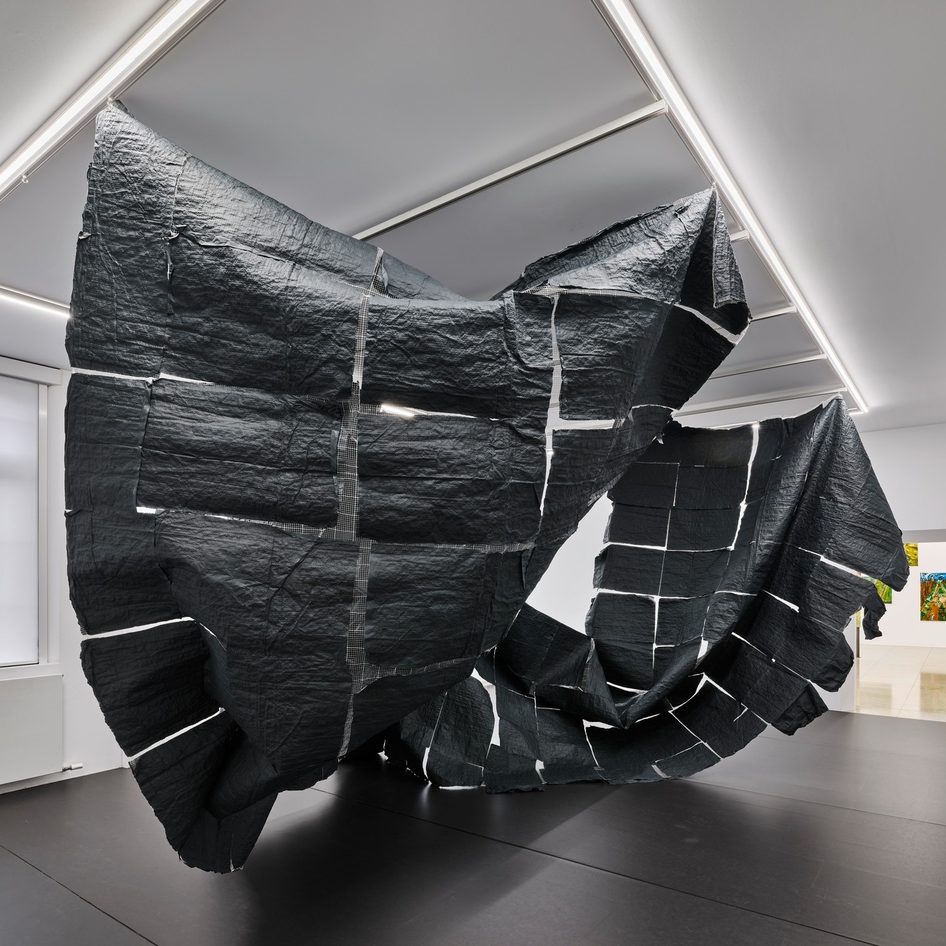 Dieser Alternativtext wurde KI-generiert: Ein großformatiges, abstraktes Objekt aus dunklem Material hängt von der Decke. Es befindet sich in einem Raum mit dunklem Boden und heller Beleuchtung.