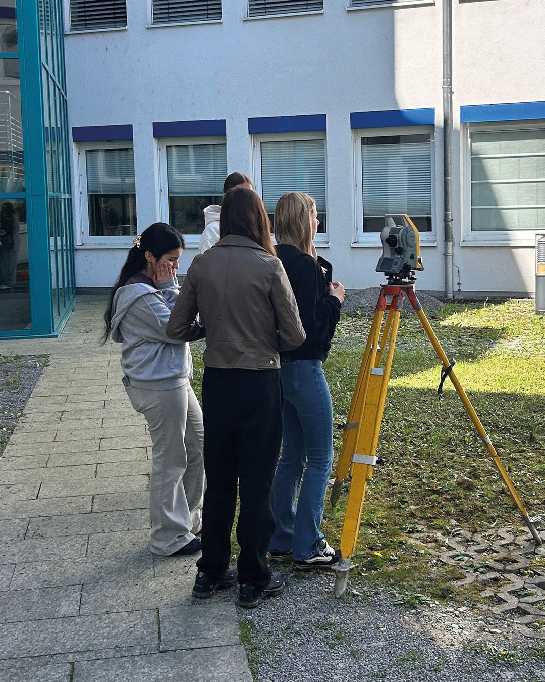Dieser Alternativtext wurde KI-generiert: Mehrere Personen stehen im Freien vor einem Gebäude zusammen und betrachten ein Messgerät auf einem Stativ. Das Bild ist farbig und zeigt einen sonnigen Tag.