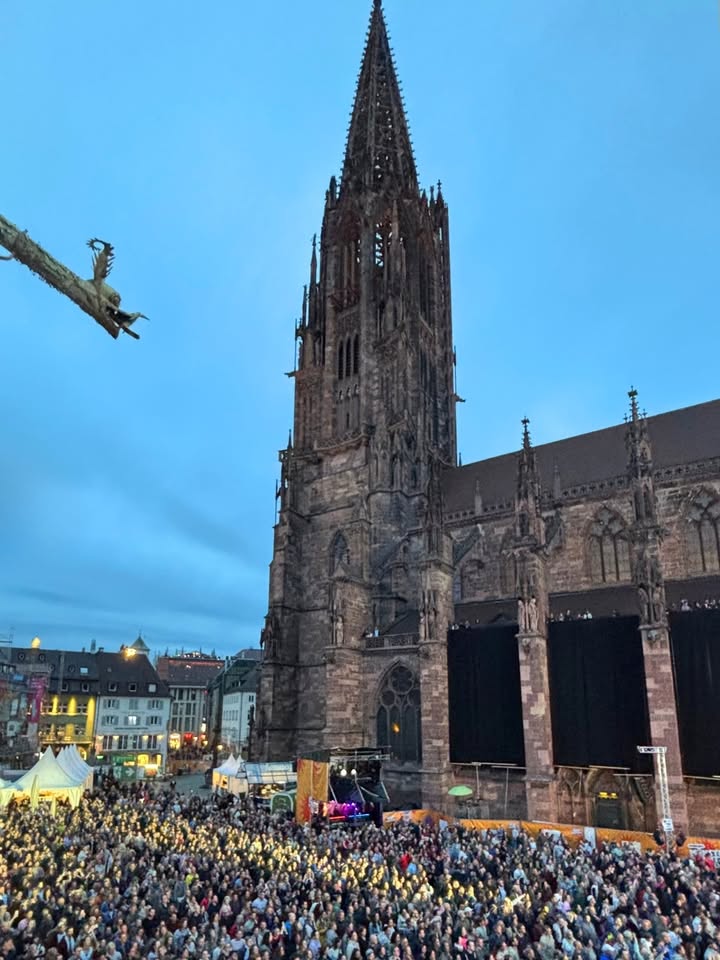 Großer gotischer Kirchturm vor blauem Abendhimmel, davor Menschenmenge auf einem beleuchteten Platz mit Bühne und Zelten, links oben Drachenfigur an einem Ast. (KI-Alternativtext)
