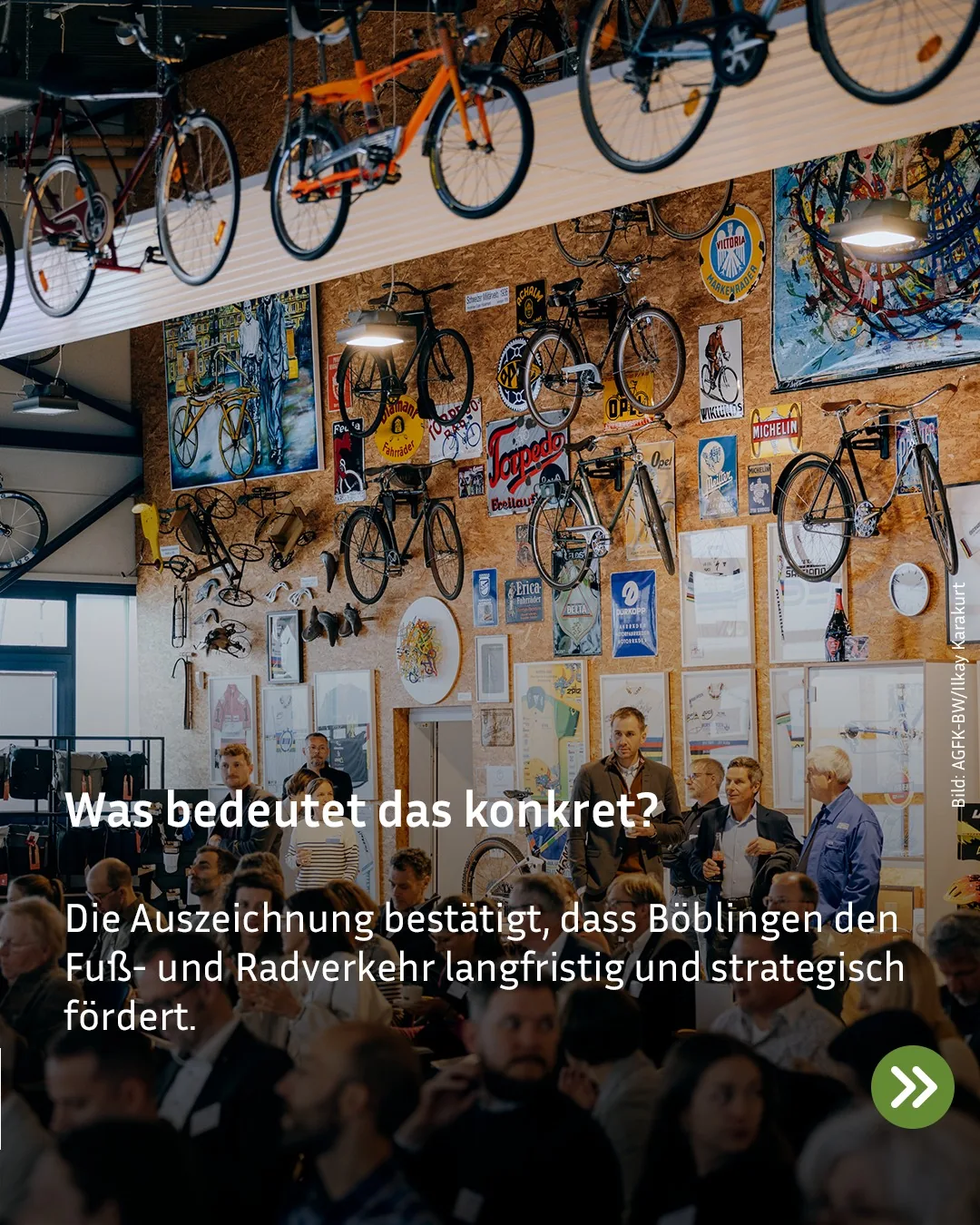 Der Raum ist mit vielen Fahrrädern an der Wand und an einer Decke sowie zahlreichen Fahrradplakaten dekoriert. Eine Menschenmenge steht in einem Veranstaltungssaal, einige Personen unterhalten sich im Vordergrund. Überlagert ist der Text "Was bedeutet das konkret? Die Auszeichnung bestätigt, dass Böblingen den Fuß- und Radverkehr langfristig und strategisch fördert." (KI-Alternativtext)