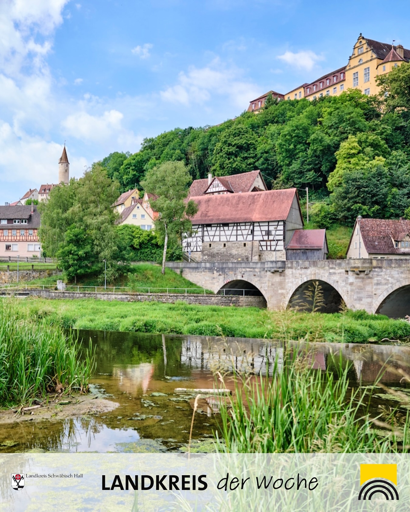 Das Bild zeigt eine Landschaft mit einem Fluss im Vordergrund, einer Brücke und Häusern im Hintergrund und einem Gebäude auf einem Hügel, alles unter einem blauen Himmel.