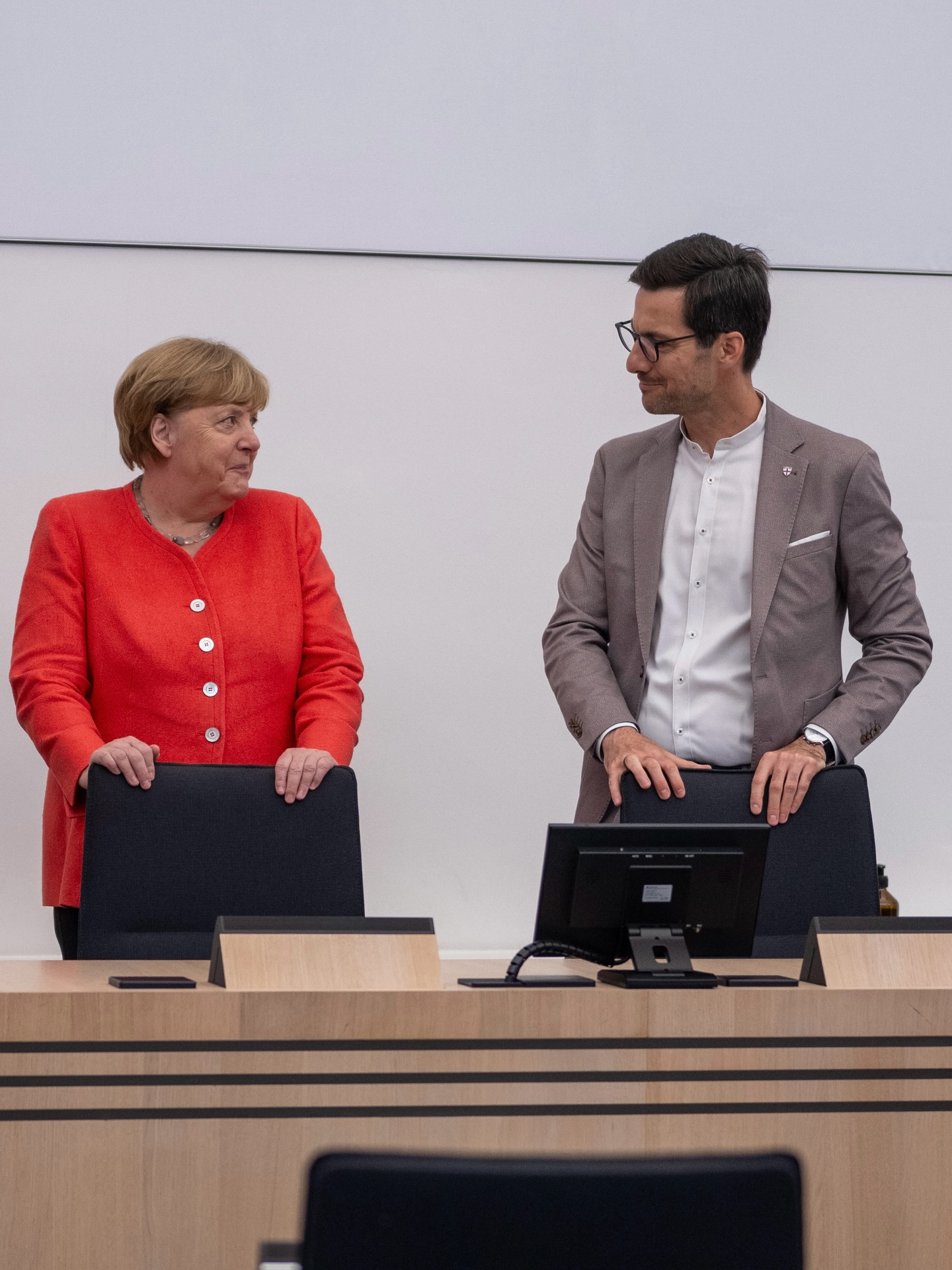 Zwei Personen stehen hinter einem Holztisch, blicken sich an und lächeln leicht. Die linke Person trägt eine rote Jacke, die rechte einen grauen Anzug mit Brille. Auf dem Tisch steht ein Monitor und ein Mikrofon. (KI-Alternativtext)