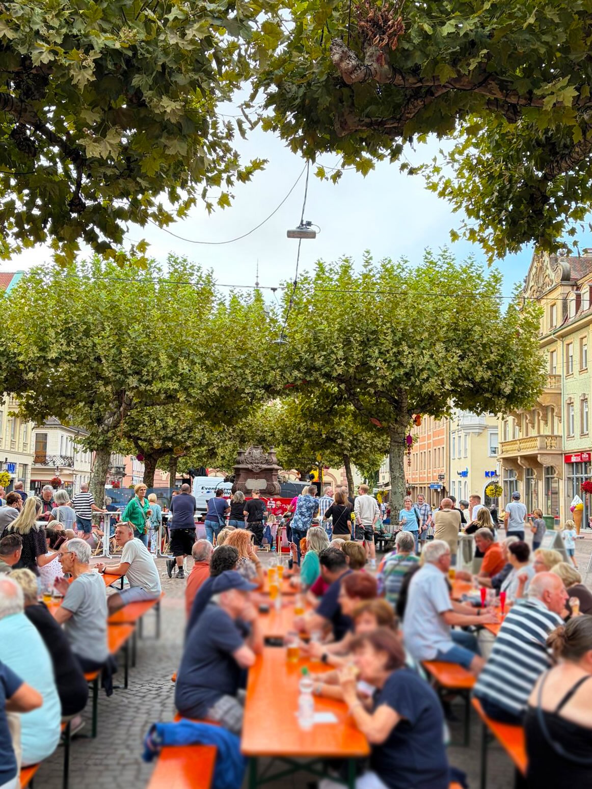 Menschen sitzen an langen, orangefarbenen Tischen auf einem Platz und essen oder trinken. Im Hintergrund stehen weitere Personen unter Bäumen vor Gebäuden, während oben Äste das Bild einrahmen. (KI-Alternativtext)