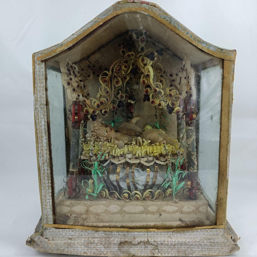 Dieser Alternativtext wurde KI-generiert: Eine dekorative Szene ist hinter Glas in einem alten Schaukasten dargestellt. Die Szene besteht aus aufwendig geformten Papierrollen und anderen handwerklichen Elementen.