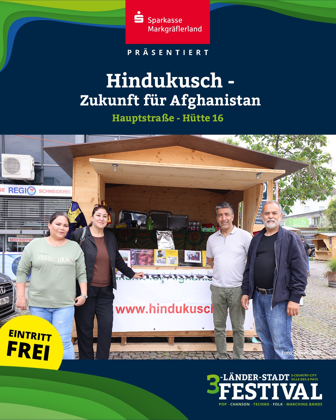 Ein Foto zeigt mehrere Personen vor einem Stand mit der Aufschrift 'www.hindukusch.de'. Im Vordergrund befindet sich ein Schild mit der Aufschrift 'Eintritt Frei'.