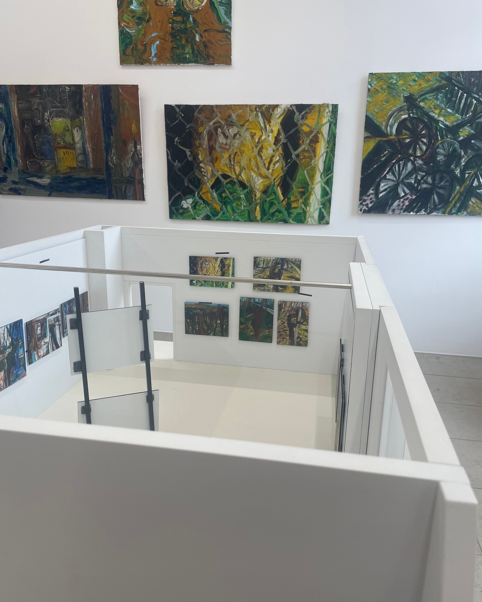 Dieser Alternativtext wurde KI-generiert: Innenansicht einer Galerie mit mehreren bunten Gemälden an den Wänden. Eine Treppe führt zu einer unteren Ebene, wo weitere Kunstwerke ausgestellt sind.