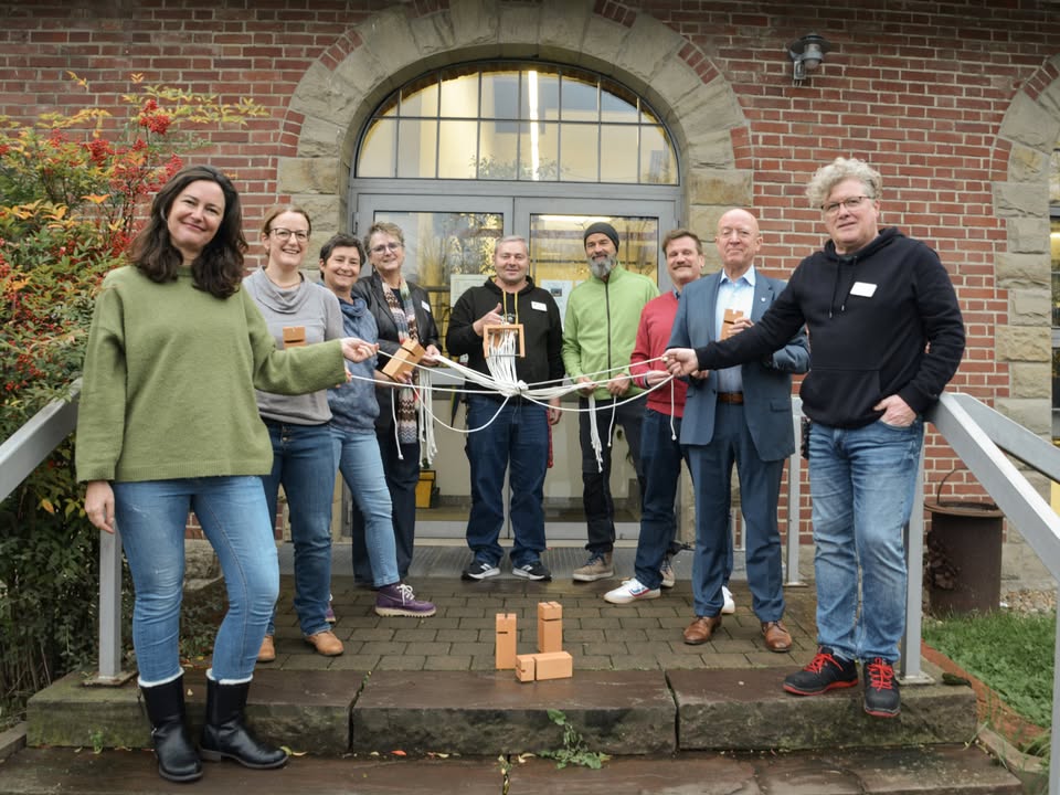 Dieser Alternativtext wurde KI-generiert: Eine Gruppe von Menschen steht gemeinsam vor einem Gebäude und hält Bänder oder Schnüre in den Händen. Sie stehen auf mehreren Stufen vor einem Hauseingang mit einem großen Bogenfenster.