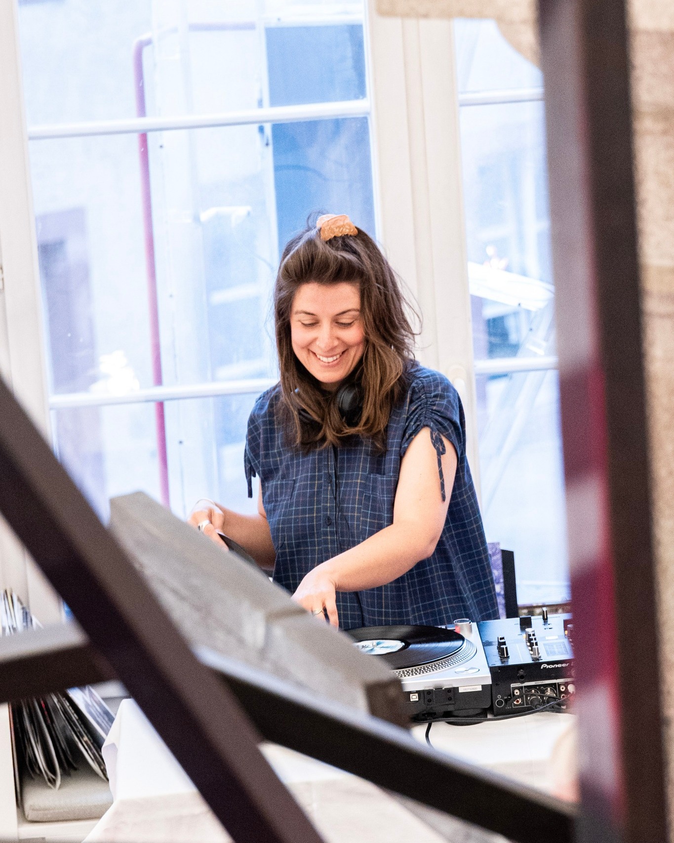 Eine Person mit langem braunen Haar und blauem kariertem Hemd lächelt, während sie an einem DJ-Setup mit Plattenspieler und Mischpult arbeitet. Kopfhörer hängen um den Hals, links daneben stehen Schallplatten in einem Korb. Im Hintergrund ist ein großes Fenster zu sehen. (KI-Alternativtext)