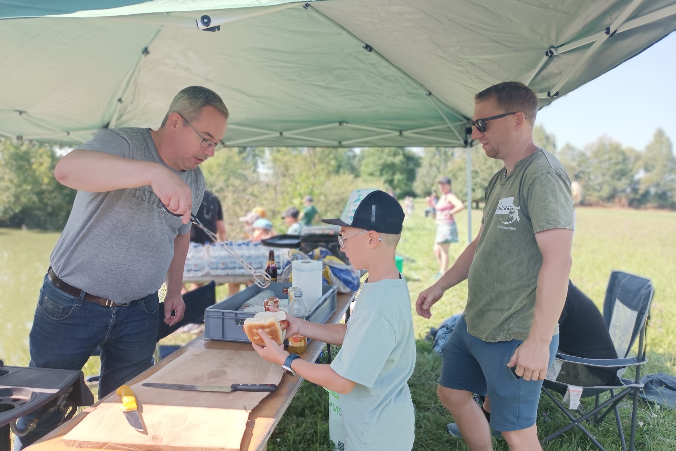 Unter einem grünen Pavillon bereitet jemand Essen zu, während andere zusehen oder sich etwas nehmen. Im Hintergrund befindet sich eine Wiese und weitere Personen.