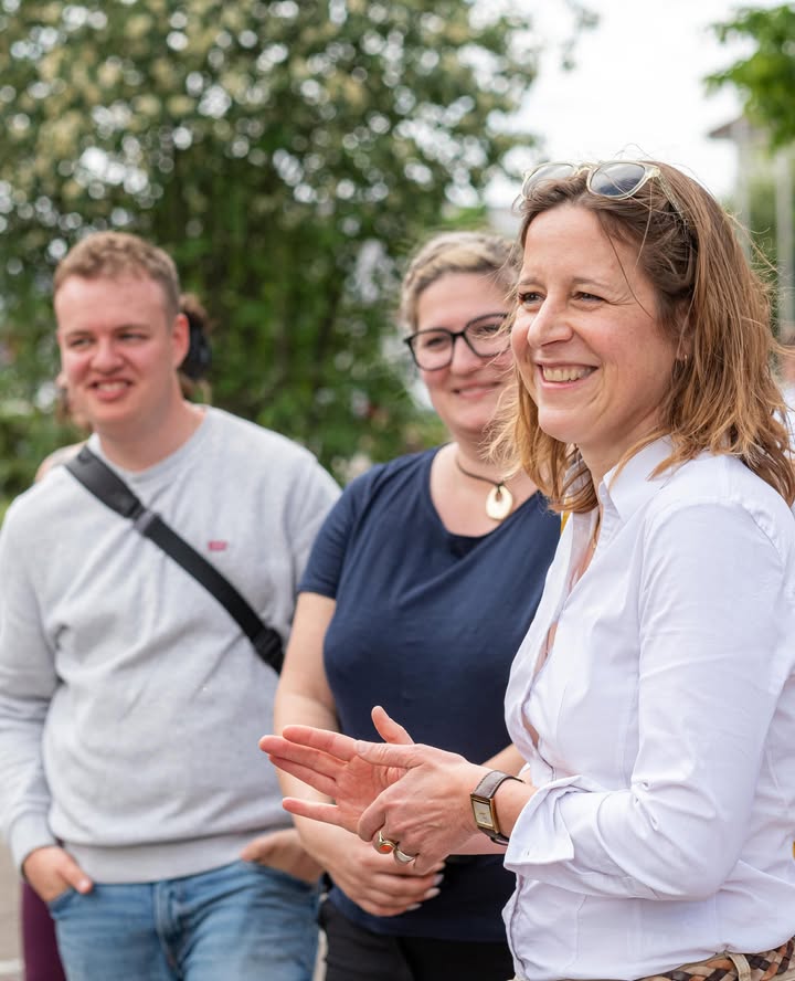 Dieser Alternativtext wurde KI-generiert: Mehrere Personen stehen im Freien vor Bäumen. Sie lächeln und wirken fröhlich, eine Person klatscht in die Hände.
